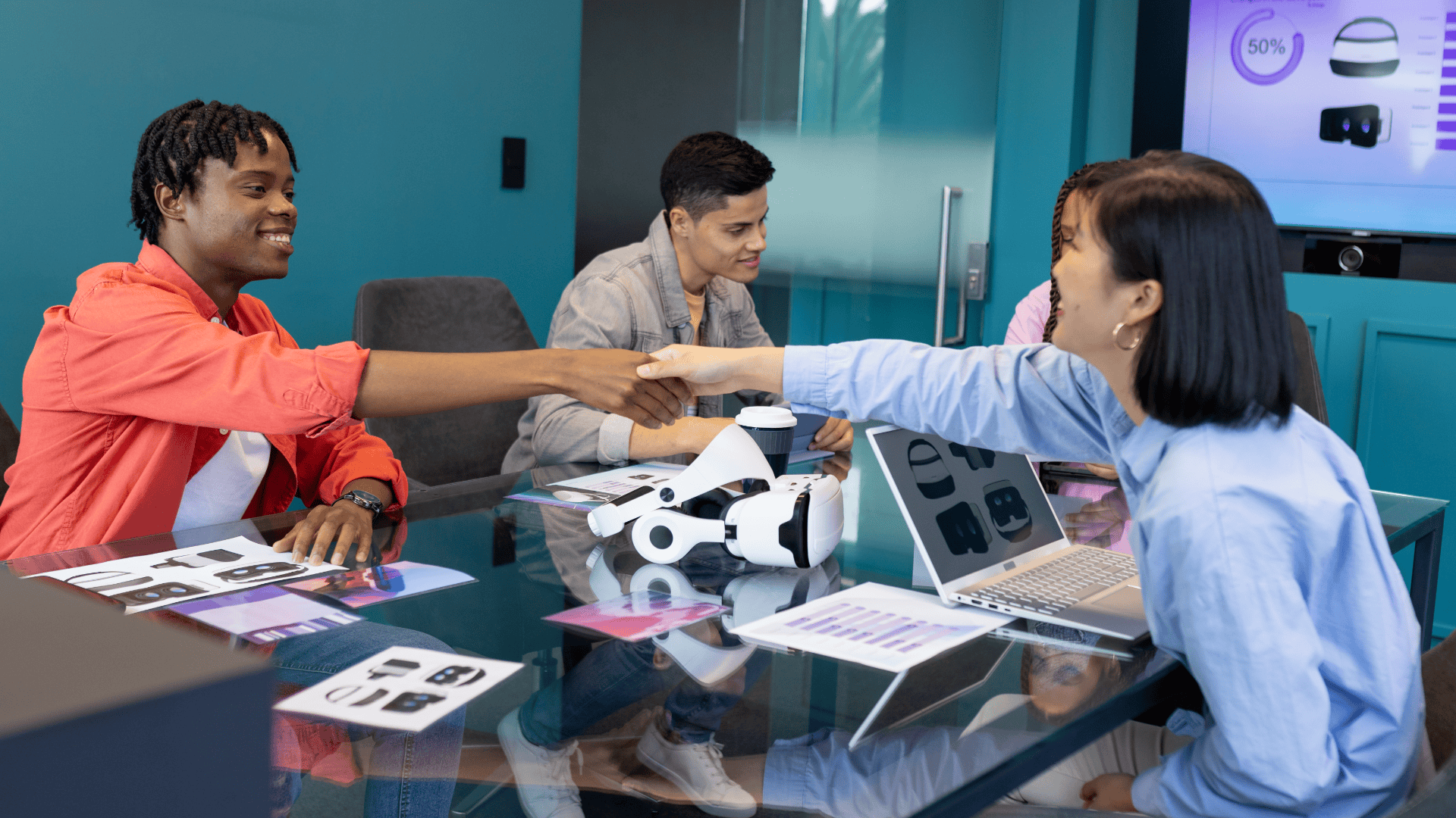 Homem e mulher jovens apertando as mãos sobre uma mesa de escritório moderna, cercados por laptops, documentos e um headset de realidade virtual (VR).