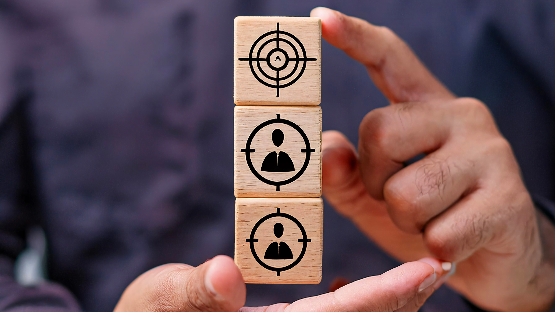 Mãos segurando uma pilha de três blocos de madeira; o topo exibe um alvo e os blocos abaixo mostram silhuetas de pessoas dentro de miras, simbolizando foco e metas.