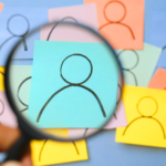 Mão segura uma lupa ampliando um desenho de figura humana em um post-it azul, destacado entre vários outros post-its coloridos com desenhos similares em segundo plano desfocado.