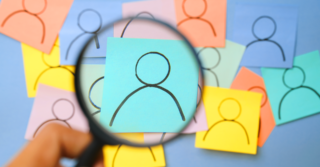 Mão segura uma lupa ampliando um desenho de figura humana em um post-it azul, destacado entre vários outros post-its coloridos com desenhos similares em segundo plano desfocado.