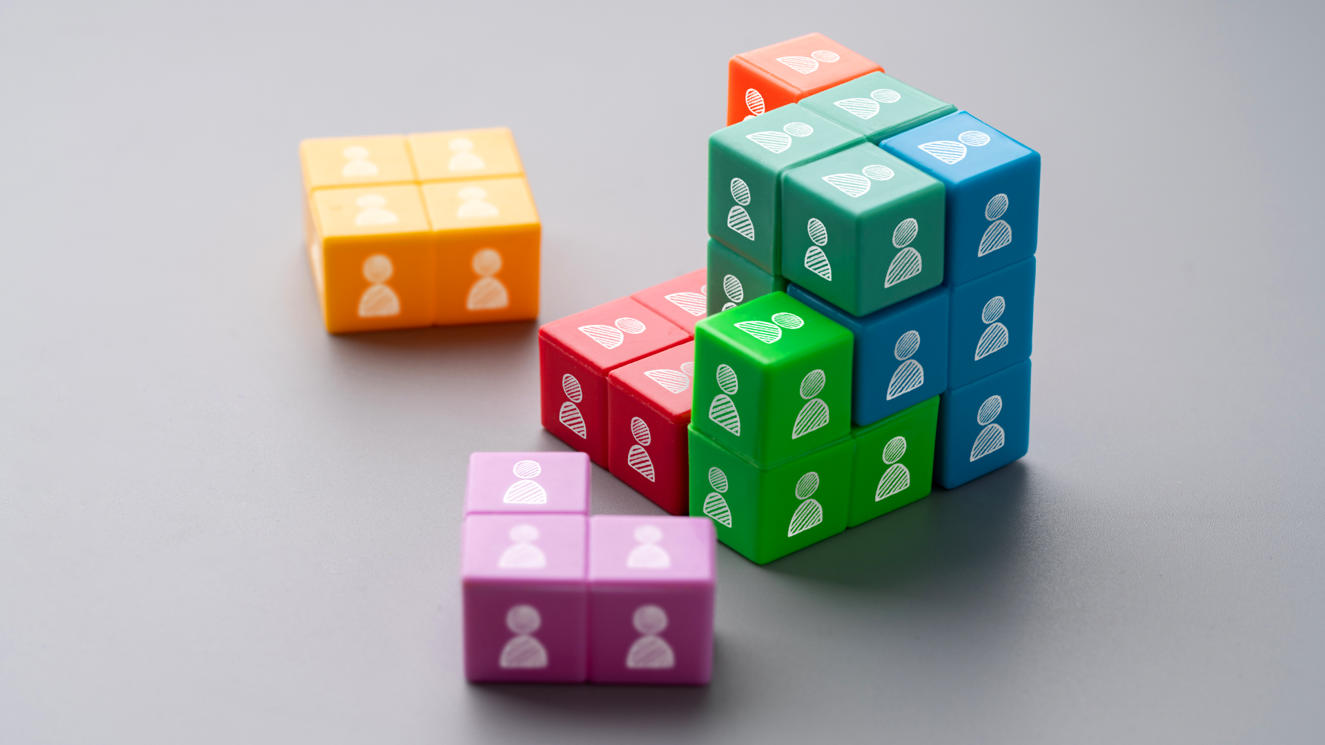 Cubos coloridos de construção com ícones de pessoas brancas estampados, sendo agrupados para formar uma estrutura sólida sobre um fundo cinza.